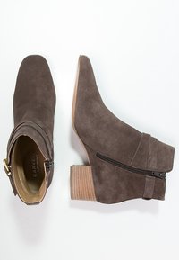 Bottes courtes en daim marron avec un bout pointu, un talon carré en bois et une fermeture éclair latérale. Caractérisées par une lanière enveloppante.