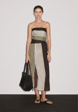 Un modèle se tient dans une robe sans bretelles multicolore avec une fente sur le côté, tenant un sac de tote en osier noir. Un fond uni met en valeur la tenue.