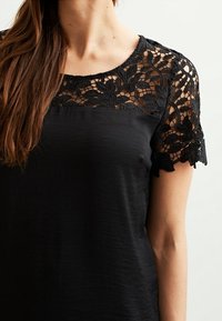 Top nero con scollatura rotonda, caratterizzato da intricate maniche in pizzo nero e un tessuto del corpo liscio, creando un contrasto di texture stratificato.