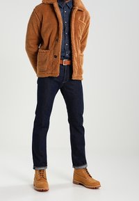 Veste brune et duveteuse avec deux poches avant, assortie à une chemise en denim. Un jeans bleu foncé et des bottes marron à lacets complètent la tenue.