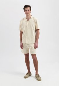 Beige overhemd met korte mouwen en bijpassende shorts van textuurstof, met een kraag en knoopsluiting. Gecombineerd met lichtbruine schoenen.