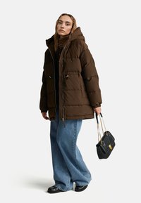 Hnědá péřová bunda s kapucí, se zipovým uzávěrem a předními kapsami. Kompletní s širokými džíny a černou kabelkou.