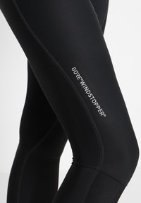 Svarta leggings med GORE® WINDSTOPPER®-logotyp, slät textur, åtsittande passform och kontrasterande sömmar längs blixtlåsen.