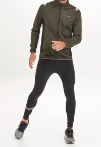 Giacca sportiva verde oliva con dettagli neri e strisce riflettenti, abbinata a leggings neri attillati e scarpe da corsa grigie.