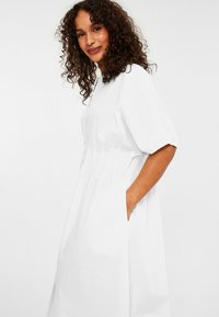 White textured dress with short sleeves, featuring side pockets and a relaxed fit, shown on a person from the side against a plain background.