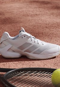 Witte sportschoen rust op een gravel tennisbaan naast een zwarte tennisracket en een gele tennisbal.