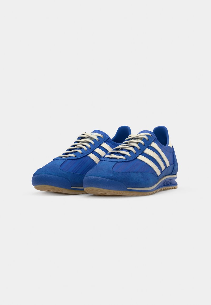Niebieskie sneakersy z białymi paskami, wykonane z zamszu i siateczki, z zaokrąglonym noskiem, płaskimi sznurowadłami oraz gumową podeszwą.