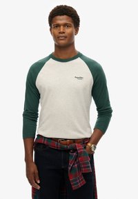 Koszula z długim rękawem typu raglan z szarym korpusem, zielonymi rękawami i małym logo. Noszona z czerwoną i zieloną kraciastą koszulą przewiązaną w talii.