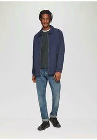 Marine blauwe zip-up jas over een donkergroene trui, gecombineerd met lichte jeans en zwarte sneakers. Soepele stof, ontspannen pasvorm, opgerolde mouwen.