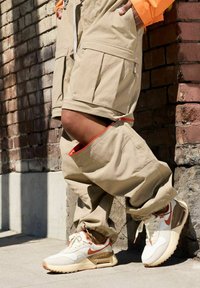 Pantaloni cargo beige con grandi tasche e dettagli della cerniera interna arancione, abbinati a sneaker Nike Air Max bianche e arancioni.