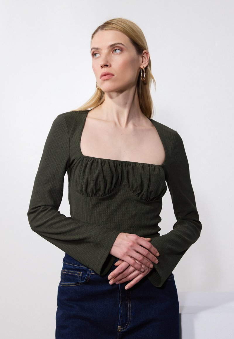 Mørkegrønn langermet topp med firkantet halslinning og rynket frontdetalj. Stoffer har en teksturert avslutning. Båret med et denimskjørt.