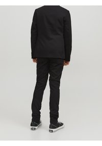 Svart kavaj med modern passform, kombinerad med svarta smala byxor. Skorna är svarta med vita detaljer. Neutralt bakgrund.