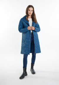 Quiltet blå frakke med hætte, trykknap- og lynlåslukning, sidelommer, parret med mørke jeans og sorte ankelstøvler. Glat tekstur, middellang design.