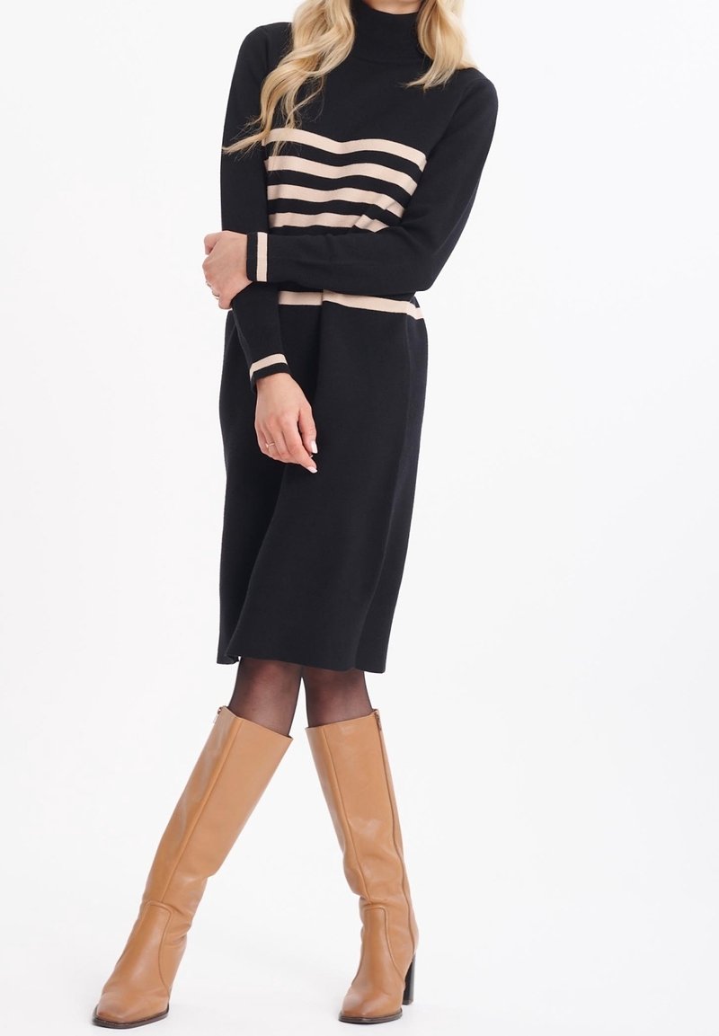 Kvinna i en svart midiklänning med beige horisontella ränder och bruna knähöga klackstövlar, som står mot en vit bakgrund.