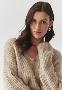 Beige gebreide trui met een V-hals en ribbelstructuur, met losse mouwen. Het model heeft een gouden creooloorbel en is zacht verlicht.
