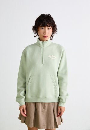 Sweatshirt en polaire vert clair avec un col zippé, doté d'une poche sur le devant et d'un texte brodé sur la poitrine. Porter par-dessus une jupe plissée marron.