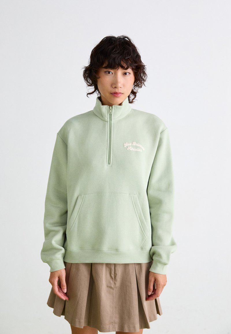 Sweatshirt en polaire vert clair avec un col zippé, doté d'une poche sur le devant et d'un texte brodé sur la poitrine. Porter par-dessus une jupe plissée marron.
