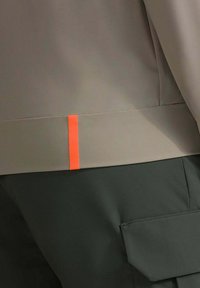 Giacca di tessuto grigio con una leggera lucentezza, con un accento arancione all'orlo. Abbinata a pantaloni verde scuro con una tasca laterale.
