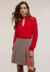 Blusa rossa con scollatura a chiave e maniche lunghe trasparenti, abbinata a una gonna a quadri con pieghe e dettagli laterali rossi.