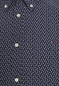 Marineblauw overhemd met knopen en een wit bladmotief, voorzien van witte plastic knopen en een klein rood accent nabij het borstzakje.