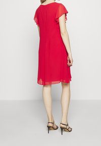 Femme portant une robe rouge longueur genou avec des manches flottantes et des sandales à talons noirs, se tenant devant un fond blanc.