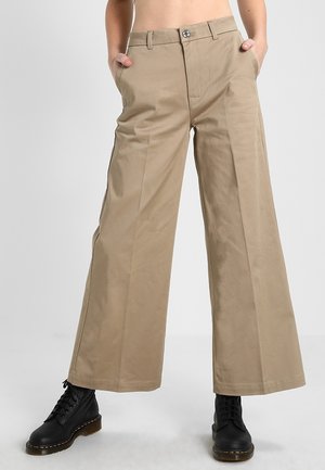 Beige wide-leg pants with front crease and side pockets, worn with black lace-up boots, model standing with hands in pockets.