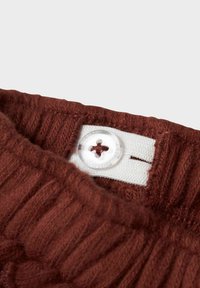 Primo piano di una cintura di un indumento lavorato a maglia marrone, dotata di una fascia elastica bianca regolabile con un bottone trasparente.