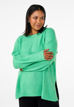 Maglione verde a maglia con maniche lunghe, texture a coste, scollatura rotonda, spacchi laterali e una vestibilità rilassata. Abbinato a pantaloni scuri.