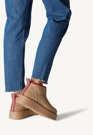 Bottines beige avec des accents rouges, en daim, dotées d'une semelle épaisse et d'un détail tressé le long du talon, associées à un jean.