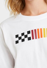 Vit sweatshirt med ett svart rutmönster på vänster sida och ett övergående mönster av röda, orange och gula linjer över bröstet.