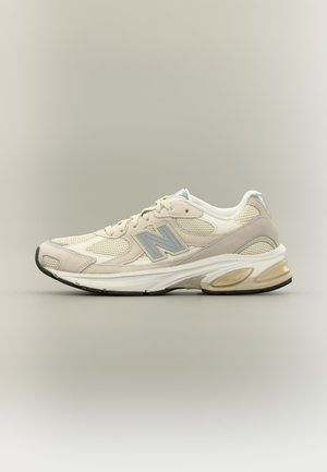 Chaussure de sport New Balance beige et blanche avec des empiècements en mesh, des surpiqûres en suède, une fermeture à lacets et une semelle rembourrée avec une unité d’air visible.