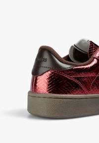 Zapatilla deportiva metálica roja con un acabado texturizado y en relieve. Presenta un talón de ante marrón y una suela de goma con un diseño de pequeñas piedras.
