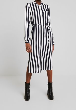 Femme portant une robe midi rayée verticale noire et blanche avec une ceinture et des bottines noires, debout devant un fond uni.