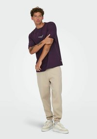 Lilla kortærmet T-shirt med logo, parret med beige sweatpants og hvide sneakers. Modellen står med armene over kors og har tatoveringer.