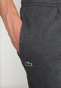 Grå bomullsbyxor med elastisk midja, sidofickor och broderad grön krokodil-logo på vänster fickområde.