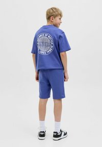 Felpa azzurra a maniche corte con una grafica del globo sul retro, abbinata a pantaloni corti blu coordinati e scarpe da ginnastica nere con dettagli bianchi.
