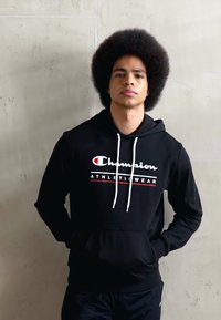 Svart hettegenser med logo foran, hvite snorer, ribbede mansjetter og hem. Modellen står mot en teksturert betongbakgrunn.