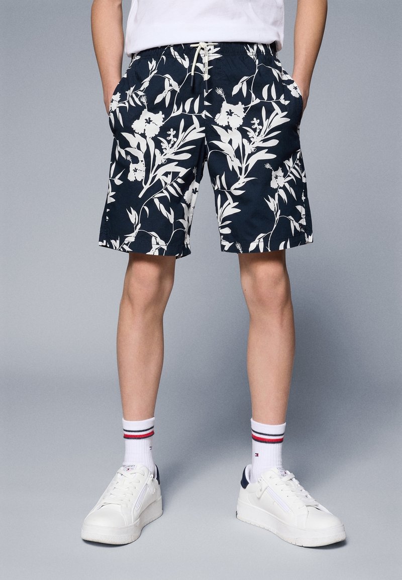 Shorts navy à motif floral avec des feuilles blanches, taille élastique, poches latérales, associés à des baskets blanches et des chaussettes rayées.