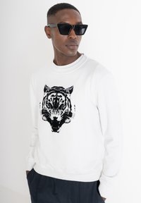 Sudadera blanca con un gráfico de tigre negro y ojos azules. Diseñada con cuello redondo y puños acanalados. Llevada con pantalones oscuros.