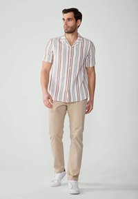 Man walking wearing short-sleeve vertical striped shirt, beige pants, and white sneakers against plain light background.