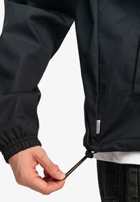 Chaqueta negra y ligera con puños elásticos y cordón ajustable en el dobladillo. Presenta una etiqueta en la costura lateral.