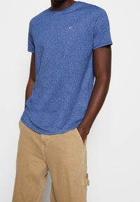 Camiseta de algodón azul jaspeada de manga corta con cuello redondo y un pequeño logo en el pecho, combinada con pantalones claros en color caqui.