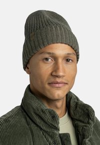 Ribbed green beanie with a small brand patch. Worn with a textured green corduroy jacket and a light olive shirt underneath.