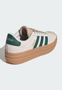 Sneakers Adidas beige e verdi con tomaia in suede, tre strisce verdi e suola in gomma testurizzata. Sono visibili dettagli di branding in oro.