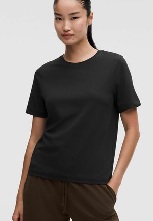 Mujer que lleva una camiseta negra lisa de manga corta y pantalones marrones con una mano en el bolsillo sobre un fondo liso.