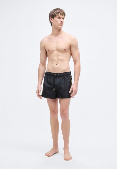 Μαύρα μαγιό σορτς με σφιχτή μέση με κορδόνι, διακοσμημένα με χρυσές λεπτομέρειες και λογότυπο στο πλάι. Κατασκευασμένα από ελαφρύ, απαλό υλικό.