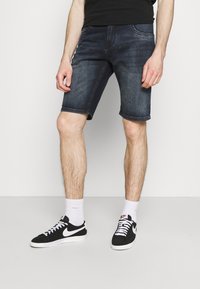 Džínové šortky tmavě modré barvy s klasickým pěti kapsovým designem, ohrnutým lemem a hladkou texturou. Kombinované s černými teniskami a bílými ponožkami.