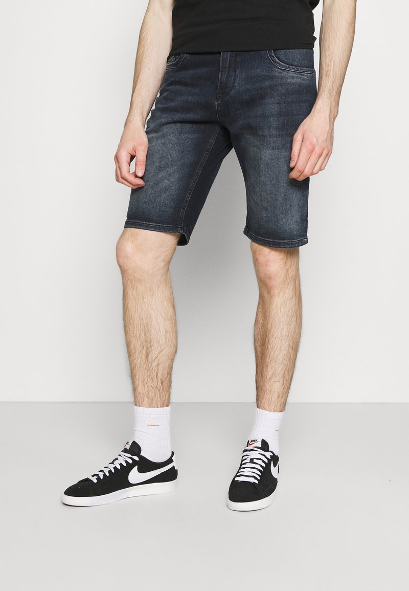 Džínové šortky tmavě modré barvy s klasickým pěti kapsovým designem, ohrnutým lemem a hladkou texturou. Kombinované s černými teniskami a bílými ponožkami.