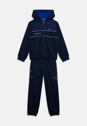 Tmavomodrý dvojdielny teplákový komplet s kapucňou, rozopínateľnou bundou a elastickými manžetami, s logom Lacoste a bielymi lemami.