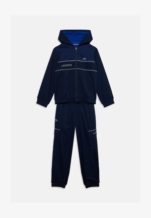 Survêtement bleu marine deux pièces avec veste zippée à capuche et manches élastiques, avec logo Lacoste et détails en passepoil blanc.